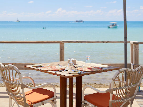 A seaside café setup featuring a table with two chairs under a large umbrella, overlooking calm ocean waters with boats in the distance.