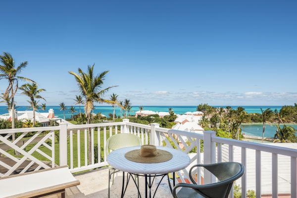 A seaside view from a balcony with a table, two chairs, and a hat on the table. The background features palm trees and a calm ocean.