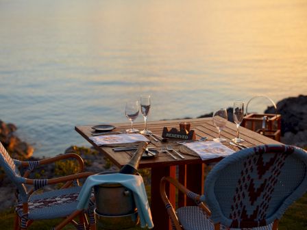 A reserved table for two set at the edge of a body of water, with menus and wine glasses, during a sunset.