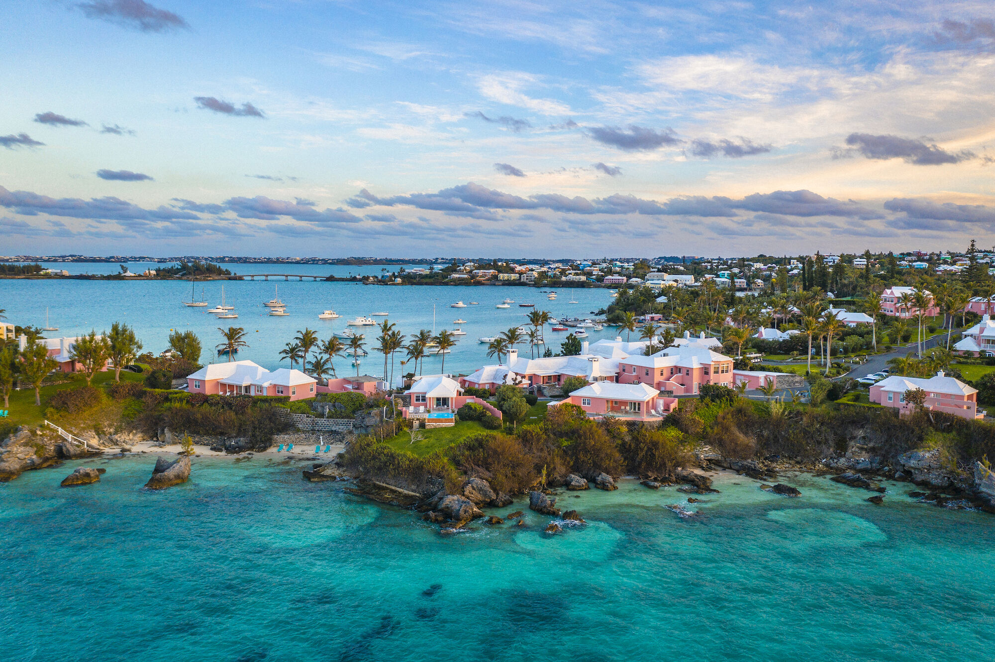 Coastal scene with pink houses, lush greenery, turquoise waters, small beach, and boats on a calm bay under a partly cloudy sky.
