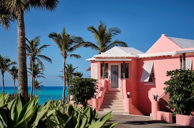A pink coastal house with stairs, palm trees, and a blue sky by the sea, sunny and tropical, inviting a relaxed island vibe.