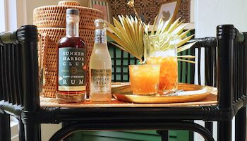 A tropical drink with a pineapple garnish sits on a black wicker chair table, flanked by rum bottles and a wicker basket.