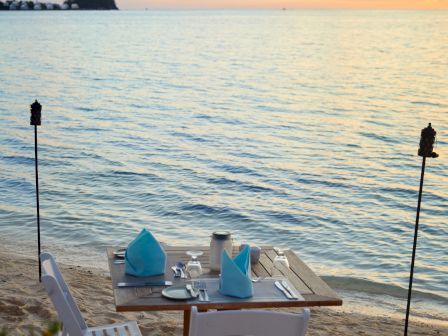 A beachside dining setup with two chairs, a table set with blue napkins, cutlery, and torches, against a backdrop of a serene ocean and sunset.