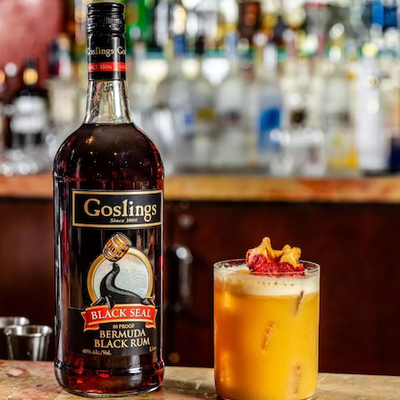 A bottle of Goslings Black Seal Bermuda Black Rum beside a tropical orange cocktail topped with fruit, on a bar counter.