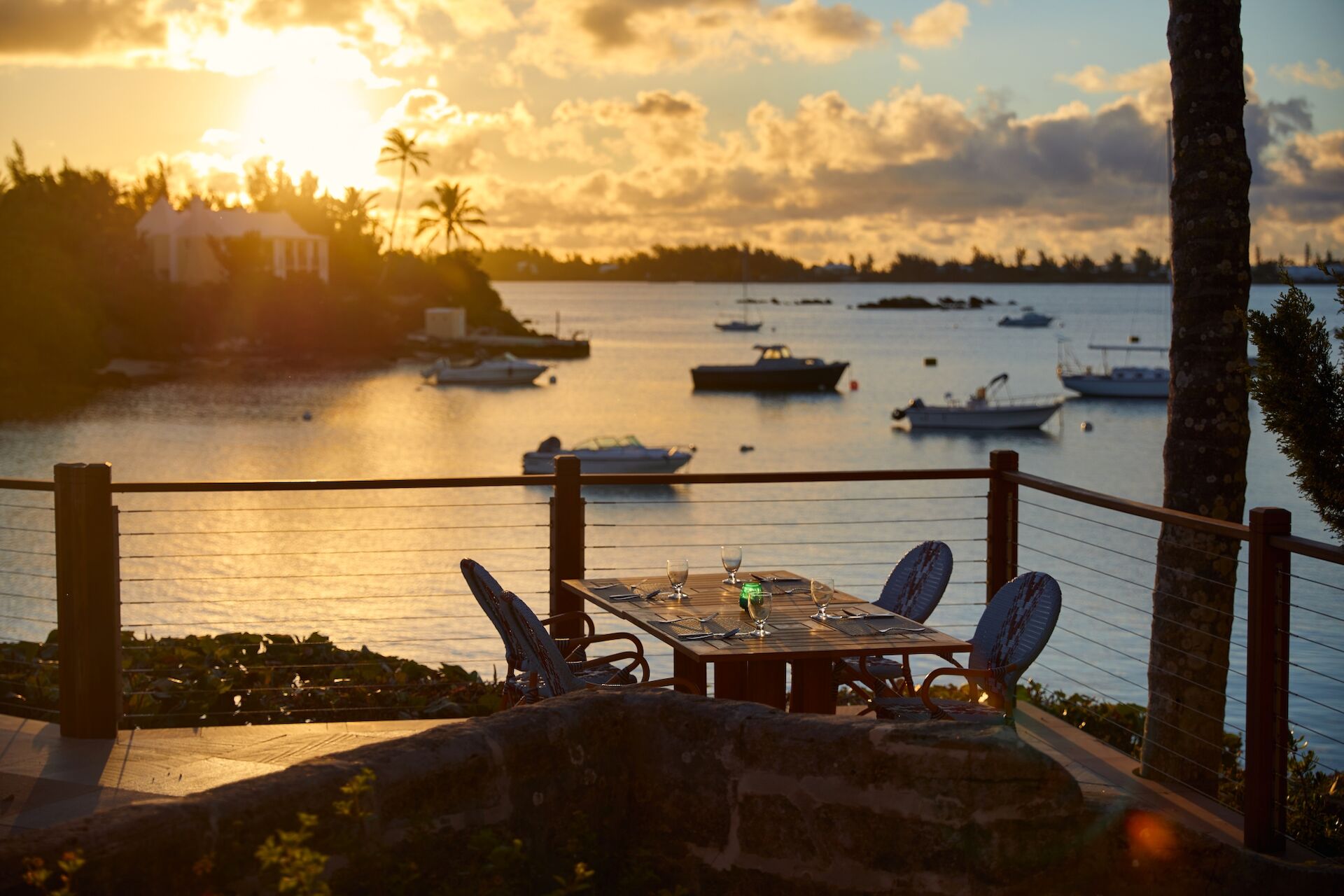 Sunset seaside view: a balcony dining table with chairs, glasses, and boats on calm water beyond.