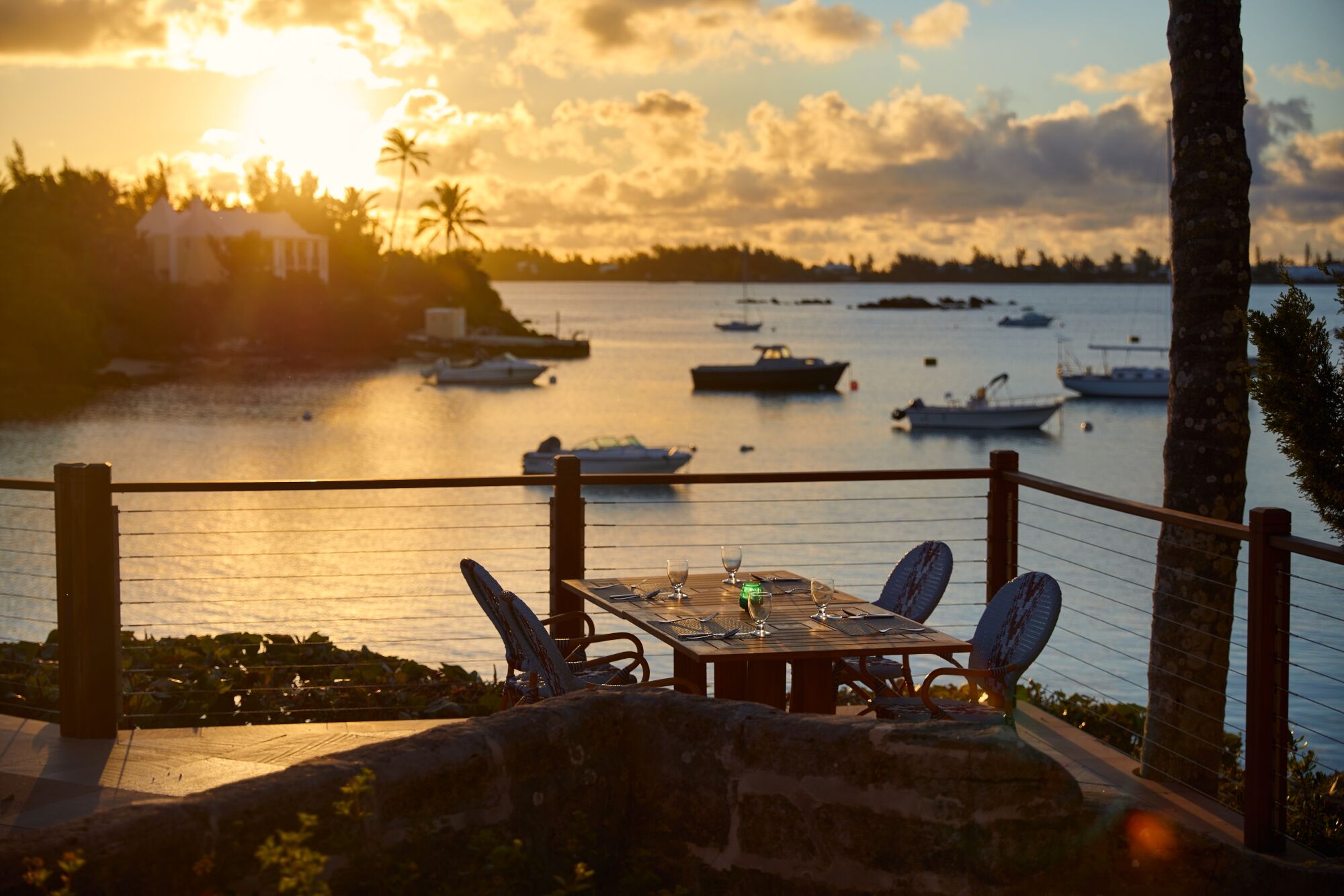 Sunset seaside view: a balcony dining table with chairs, glasses, and boats on calm water beyond.
