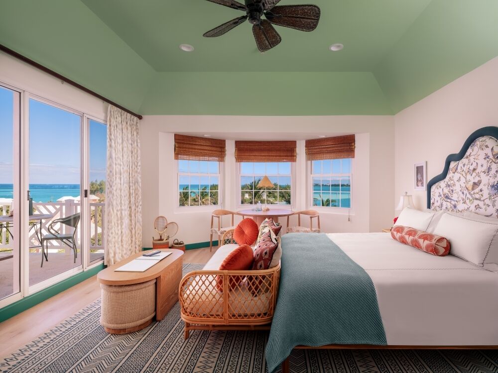 A bright tropical hotel room with a king bed, wicker seating, sea-view windows, and a balcony overlooking the ocean.