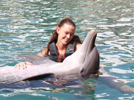 A woman in shallow water smiles while a dolphin surfaces beside her.