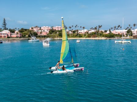 A colorful sailboat is sailing in clear blue water near a shore with pink buildings and palm trees, with other boats anchored nearby, under a blue sky.