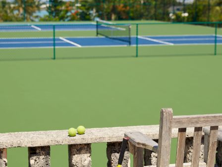 The image shows a tennis court with a bench in the foreground, a tennis racket leaning on the bench, and several tennis balls scattered nearby.