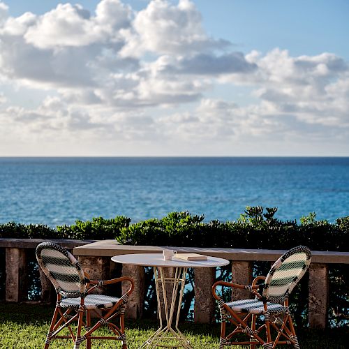Two chairs and a table are set up on grass, overlooking a scenic ocean view under a partly cloudy sky, with a stone fence and greenery in the background.