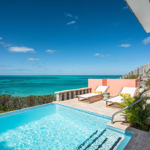This image shows a stunning beachside view with an infinity pool, two lounge chairs, clear blue skies, and a turquoise ocean, creating a relaxing scene.