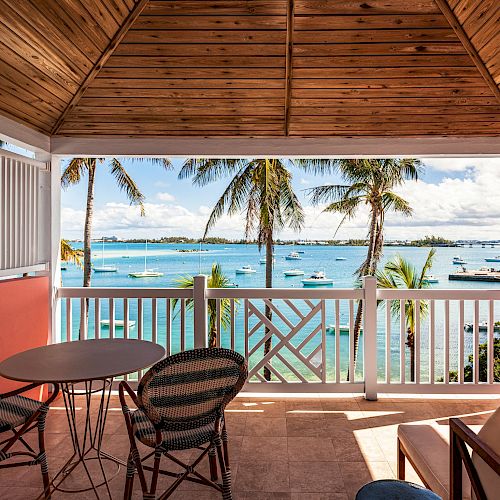 A covered balcony overlooks a tropical seascape. There are palm trees, boats on the water, an orange wall, a small table, and chairs.