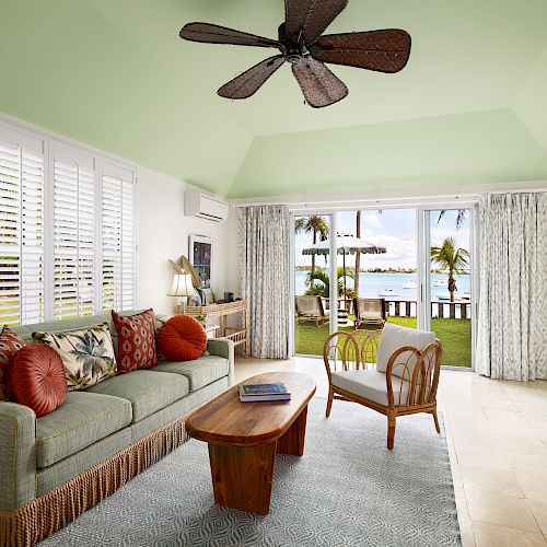 A cozy living room with green walls, wooden furniture, floral cushions, ceiling fan, and view of a grassy yard with palm trees and ocean beyond.