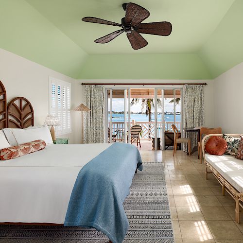 A bedroom with pastel green walls, featuring a large bed, wicker furniture, ceiling fan, and sliding doors opening to a balcony with a water view.
