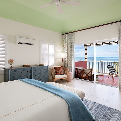 A bright, cozy bedroom with sea view features light-colored walls, a large bed, dresser, armchair, and access to a balcony with outdoor seating.