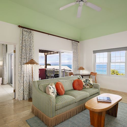 A bright living room with green walls, a sofa, pillows, a coffee table, ocean view windows, and a door leading to another room ends the sentence.