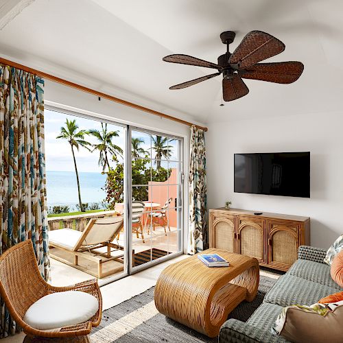 A cozy room with tropical decor features a rattan chair, sofa, wicker coffee table, and balcony with a stunning ocean view. A ceiling fan and TV are present.