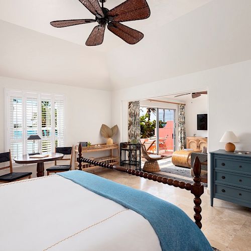This image shows a well-decorated bedroom with a ceiling fan, a bed, a dresser, a small table with chairs, and a seating area that opens to a balcony.