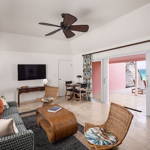 A cozy living room with wicker furniture, a TV, and an open sliding door revealing a patio with an ocean view in the background.