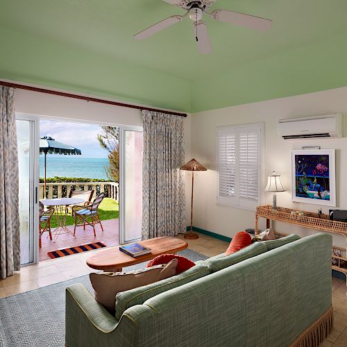 This image shows a cozy living room with a green sofa, a TV, and a desk. There is an open sliding door leading to an outdoor patio with a sea view.