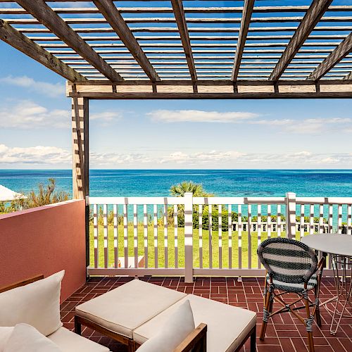 A balcony with lounge chairs and a table set is overlooking a garden and a beautiful ocean view, under a wooden pergola with a clear blue sky.