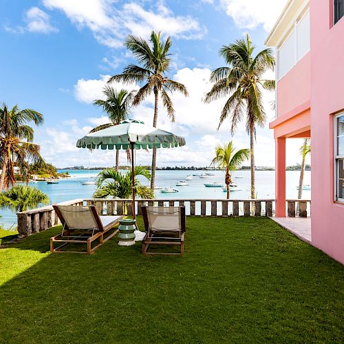 A tropical outdoor setting with palm trees, a pink building, lounge chairs, a parasol, and a scenic view of the ocean with boats in the background.