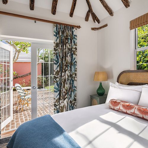 A cozy bedroom with a bed, large window, and glass doors opening to a courtyard with outdoor seating and greenery, under exposed wooden beams.