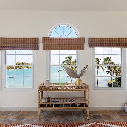 A room with three windows revealing a tropical ocean view, a wicker console table with decor, woven baskets on the side, and patterned floor rug.