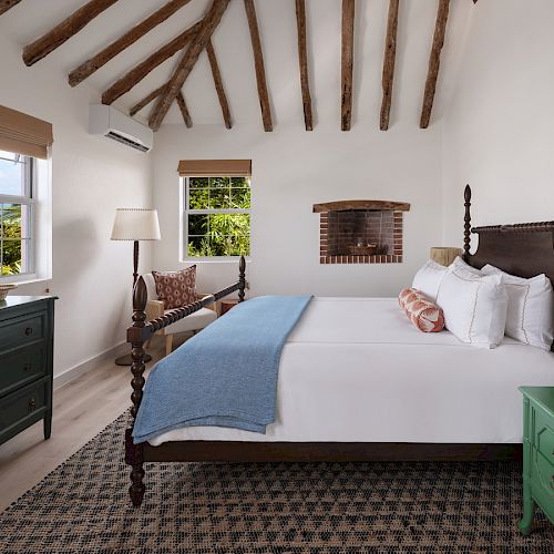 A cozy bedroom with a beamed ceiling, a large bed with a blue throw, a green dresser, and a green nightstand with lamps, and a window with a view.