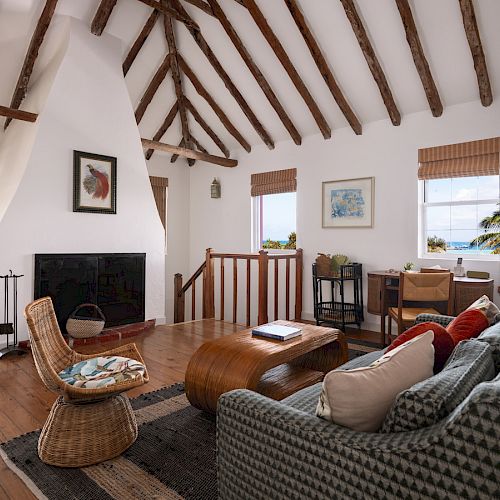 A cozy living room features a high wooden ceiling, a fireplace, a wicker chair, a cushioned sofa, a wooden coffee table, and windows with a view.