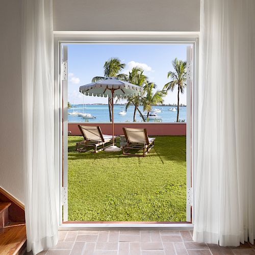 A patio view through an open door shows two lounge chairs under an umbrella, with palm trees and a coastal scene in the background.