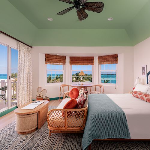 A luxurious bedroom features a large bed, cozy seating, big windows with ocean views, a ceiling fan, and access to a balcony with beach scenery.