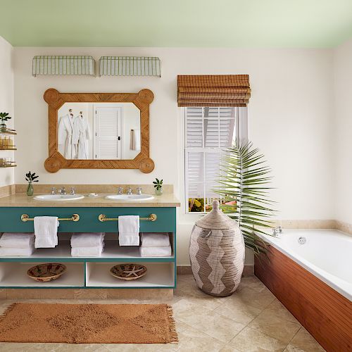 A bathroom with a double sink vanity, large mirror, bathtub, woven blinds, a floor plant, and towels stored on shelves below the sinks.
