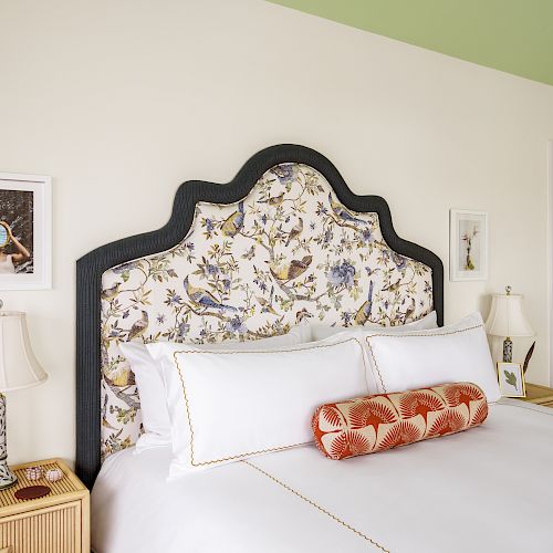 A bedroom featuring a bed with a floral headboard, white bedding, and decorative pillows. Two framed pictures, two bedside tables, and lamps complete the decor.