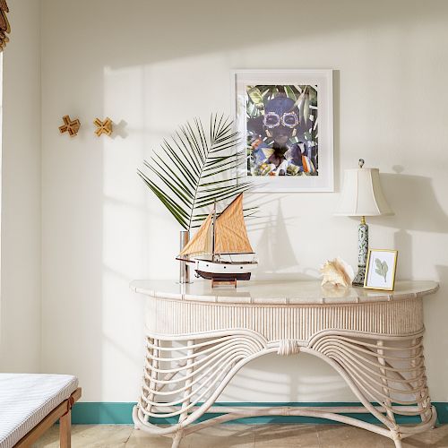 A decorative console table with a model sailboat, a palm leaf, a lamp, a shell, and a framed picture on the wall above in a sunlit room ends the sentence.