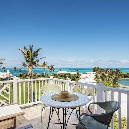 A patio overlooking a scenic ocean view with palm trees, featuring a small round table with a hat on it and two chairs.