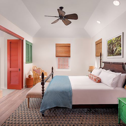 A cozy bedroom with a large bed, colorful doors, wicker chairs, a ceiling fan, and a vibrant green nightstand, all lit by warm natural light.