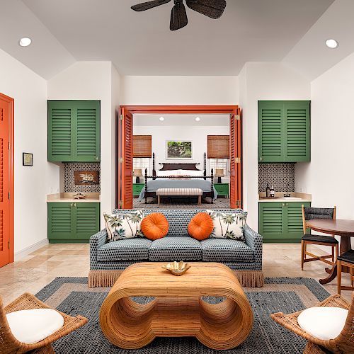 A living area with wicker furniture, a wooden table, green cabinets, an orange door, and a view into a bedroom with a bed and nightstands.