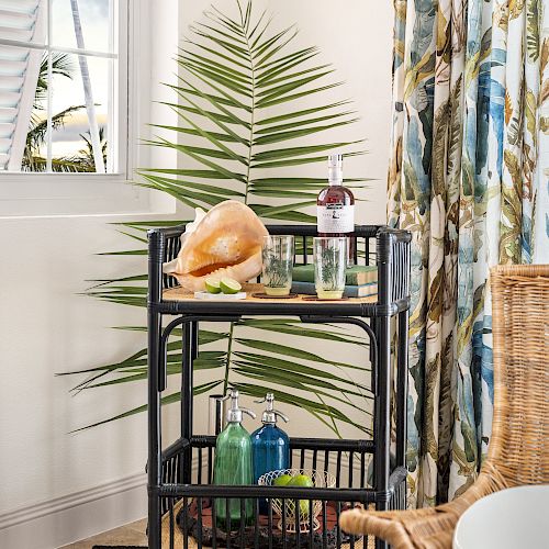 A decorative cart with bottles, a coconut, and a glass sits in front of a large leaf. There's a window, curtains, and a wicker chair in the background.