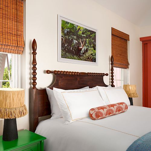 A cozy bedroom with a wooden bed, white bedding, two wicker lamps, a green nightstand, a framed nature picture, and red louvered closet doors.