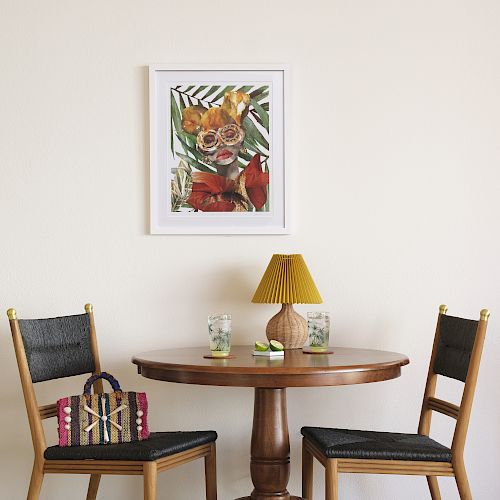 A wooden table with two chairs, a yellow lamp, two glasses, a floral picture on the wall, and something colorful on a chair.