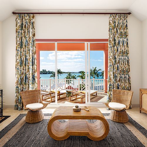 A living room with wicker furniture, tropical-themed curtains, a coffee table, and a view of the ocean through sliding glass doors ending the sentence.