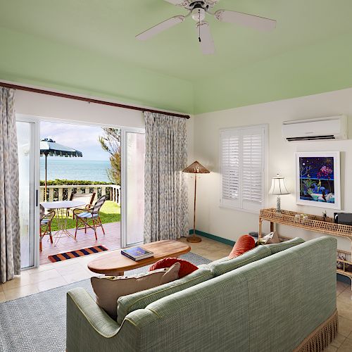A cozy living room with a green ceiling, sofa, TV, lamp, and large sliding doors opening to a patio with a sea view and greenery outside.