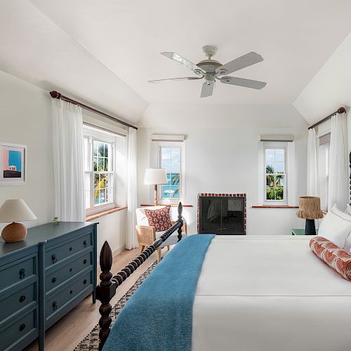 A bright bedroom with a large bed, blue dresser, ceiling fan, windows, and a seating area near a fireplace.
