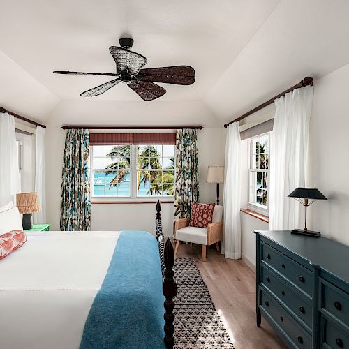 A bright bedroom with a large bed, ceiling fan, dresser, armchair, and windows displaying a tropical beach scene outside with palm trees.