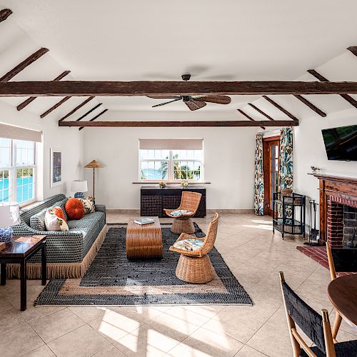 A cozy living room with exposed beams, wicker furniture, a fireplace, and large windows. Decor includes a wall-mounted TV and decorative pillows.
