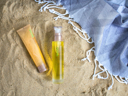 A yellow sunscreen bottle and golden tube rest on sandy beach ground with a blue towel and white edging nearby.