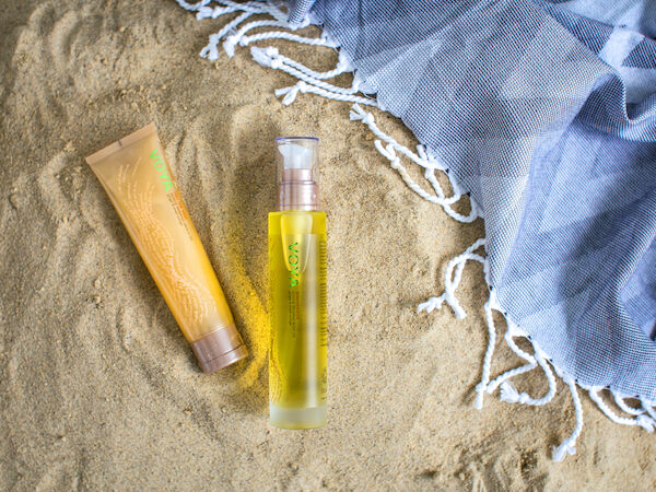 A yellow sunscreen bottle and golden tube rest on sandy beach ground with a blue towel and white edging nearby.