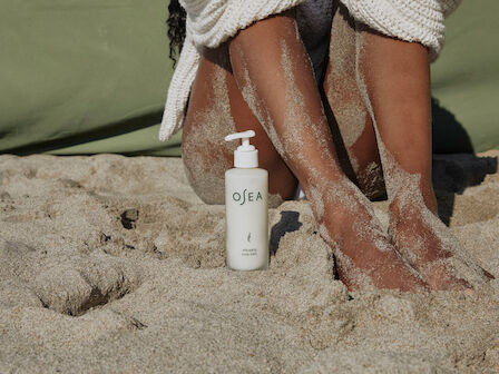 A person&rsquo;s legs in sand with an OSEA anti-aging body balm bottle on a beach blanket, suggesting skincare after sun or sand.